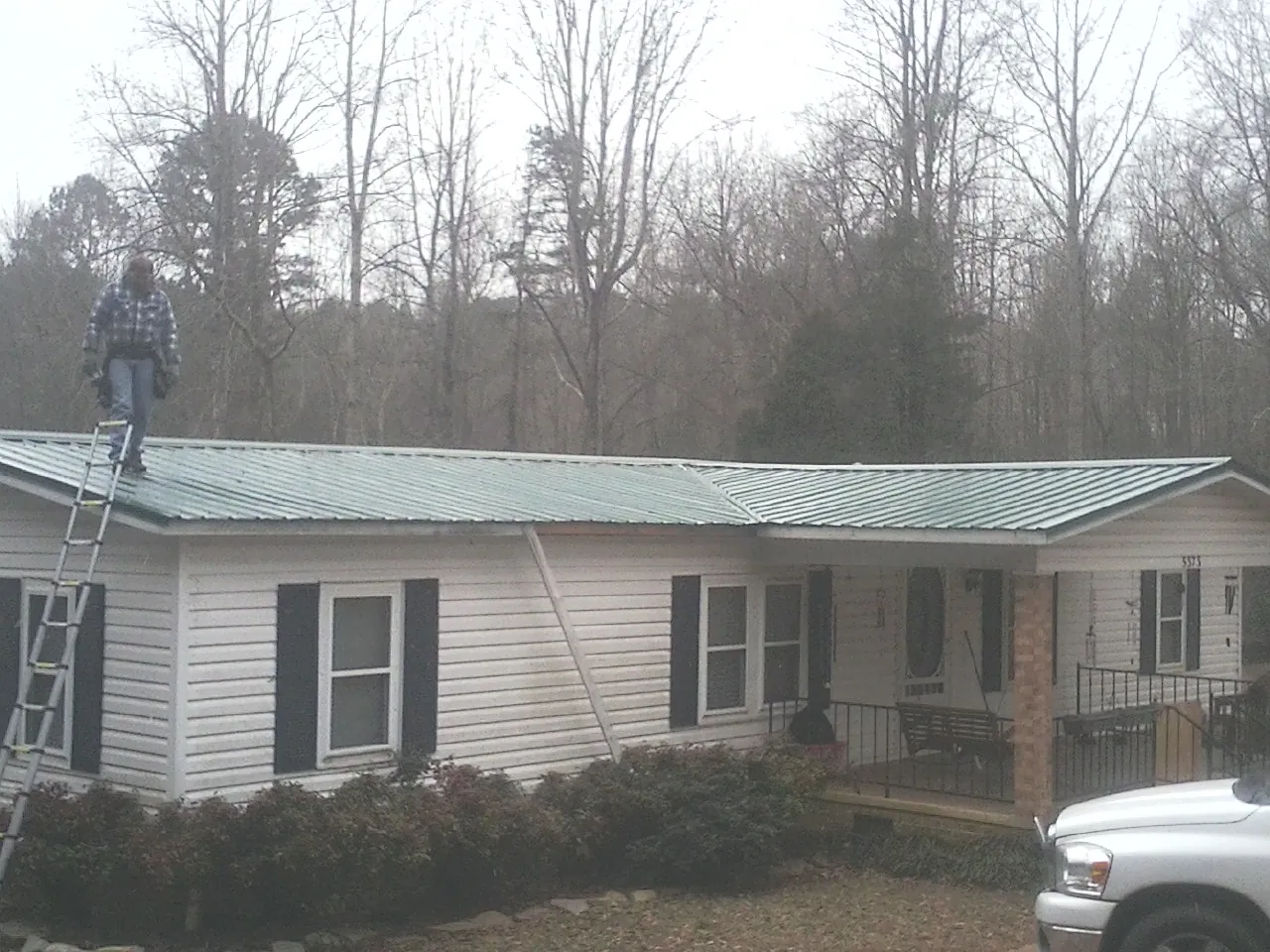 Worker on a ladder beside a new green metal roof on a ranch home in Moriches