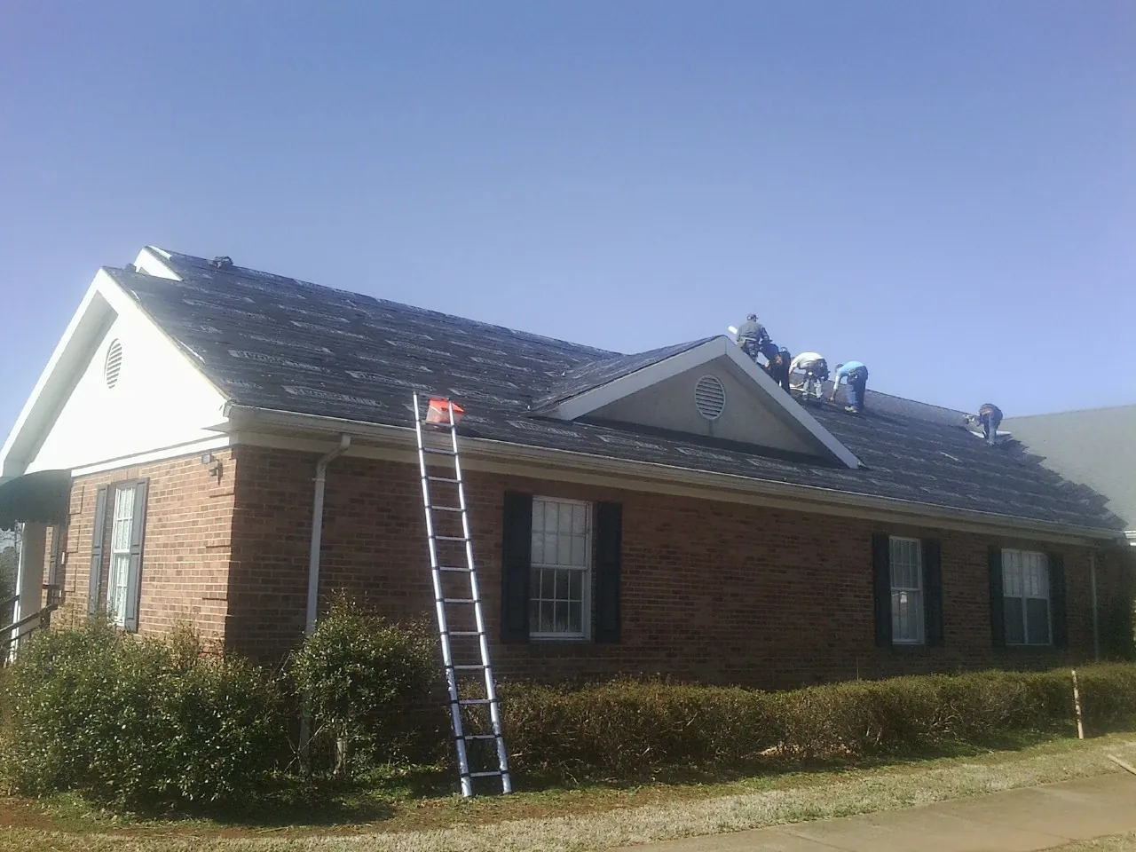 Crew tearing shingles off a brick ranch roof with a ladder set — Emergency Roof Repair work by our crew in Moriches