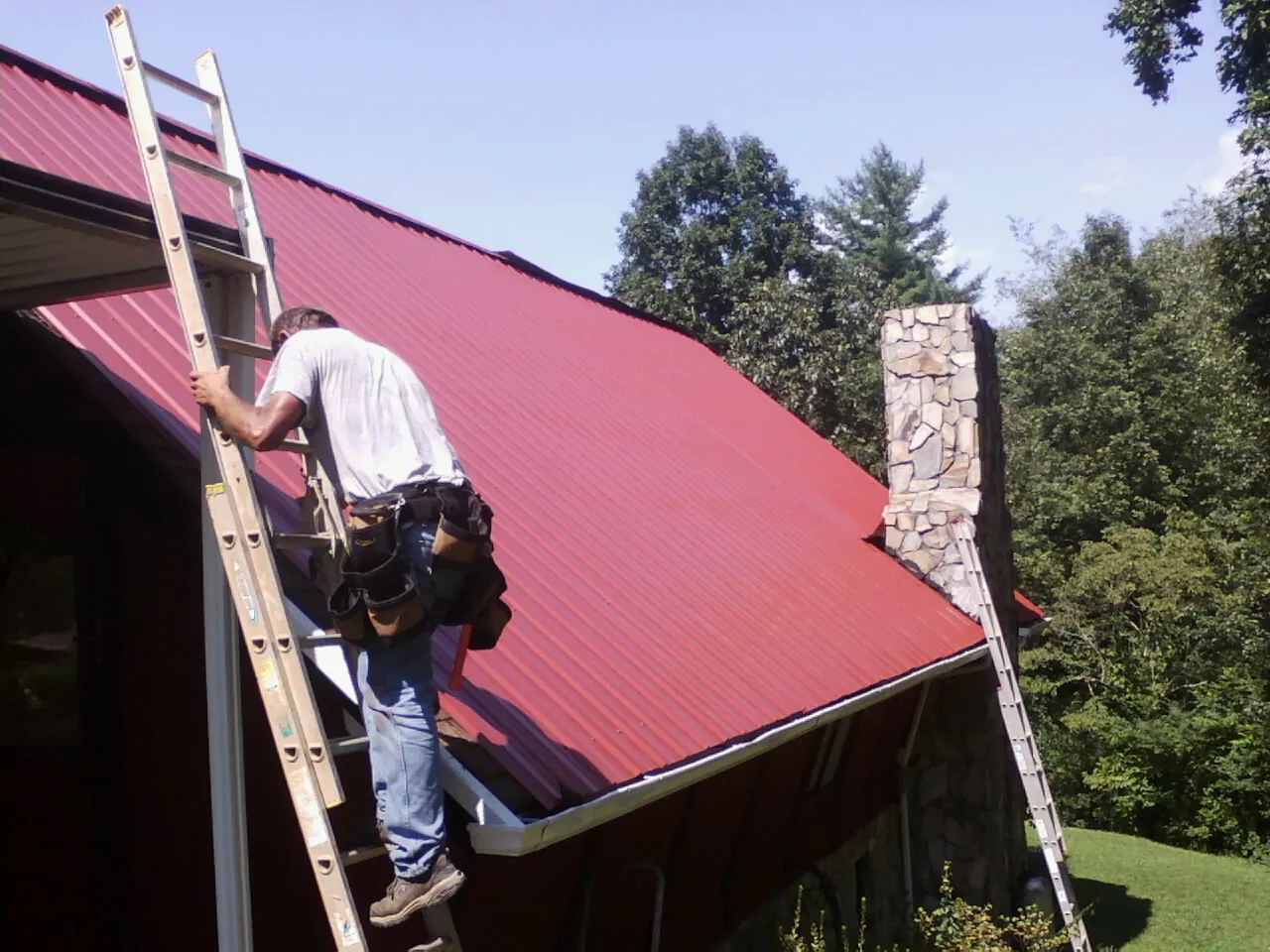 Roofer on a ladder beside a red metal roof and stone chimney — Soffit & Fascia Repair by Roofing Brothers in Moriches