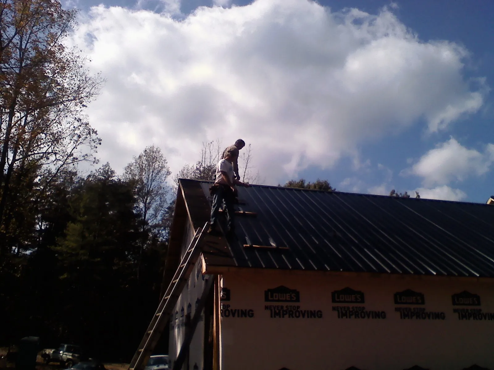 Roofer fastening dark metal panels high against a cloudy sky, from a Roof Inspection job in Moriches
